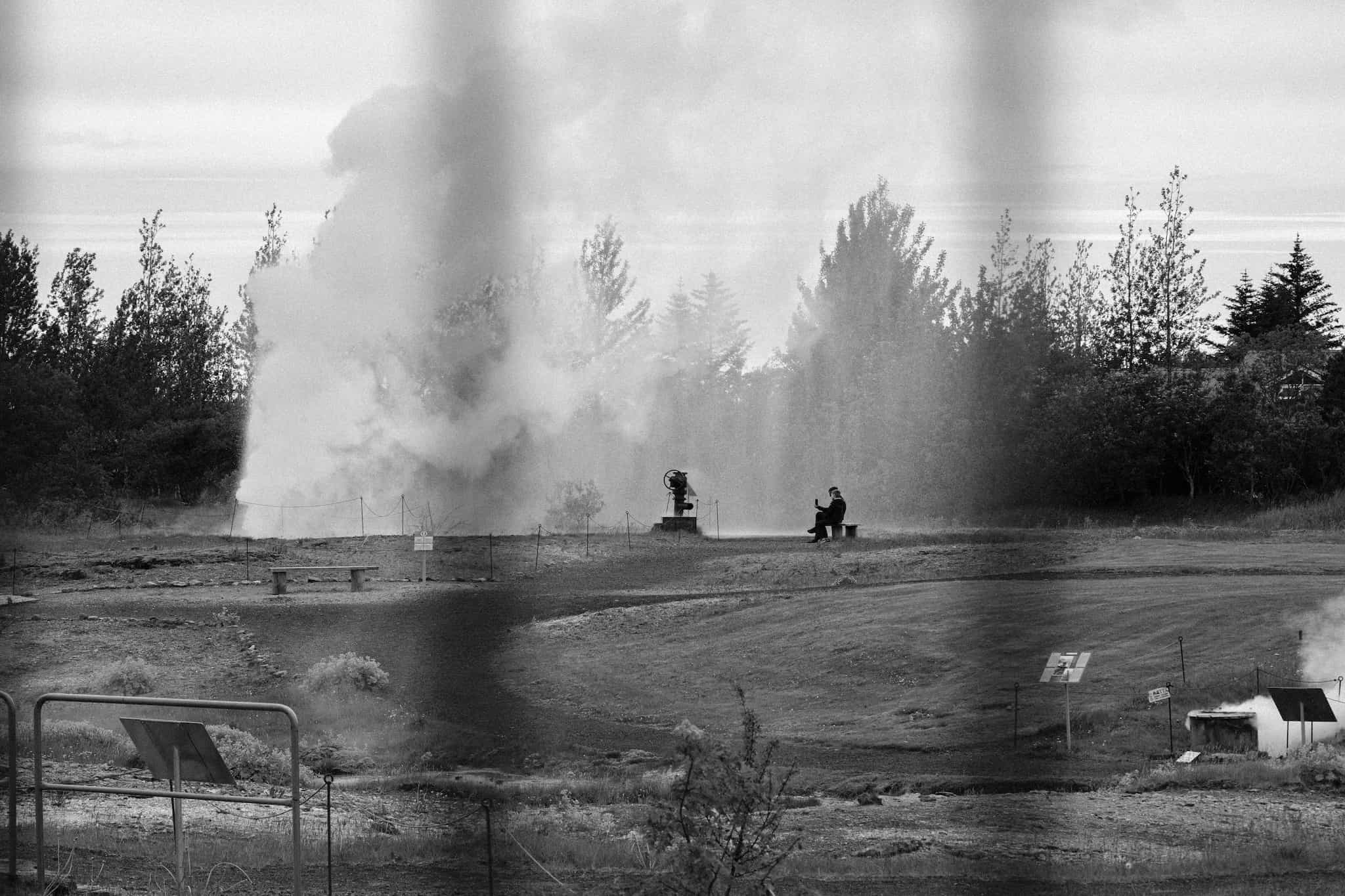 A black and white photo of a couple of tourists sitting on a bench watching a geyser erupt in the geothermal park in Hveragerði.