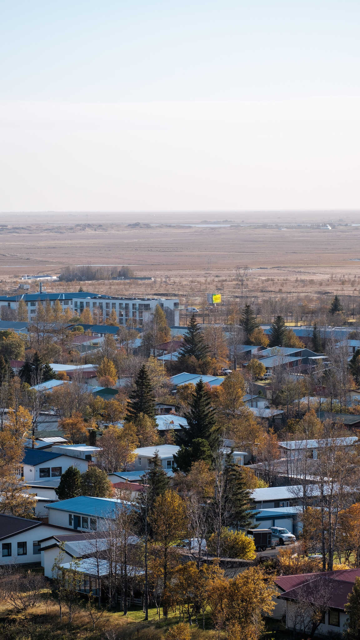 A view over Hveragerði from a hilltop. Large trees dot the landscape between low, single or two-storey, houses. In the distance you can see the town’s one large ad screen that’s currently bright yellow