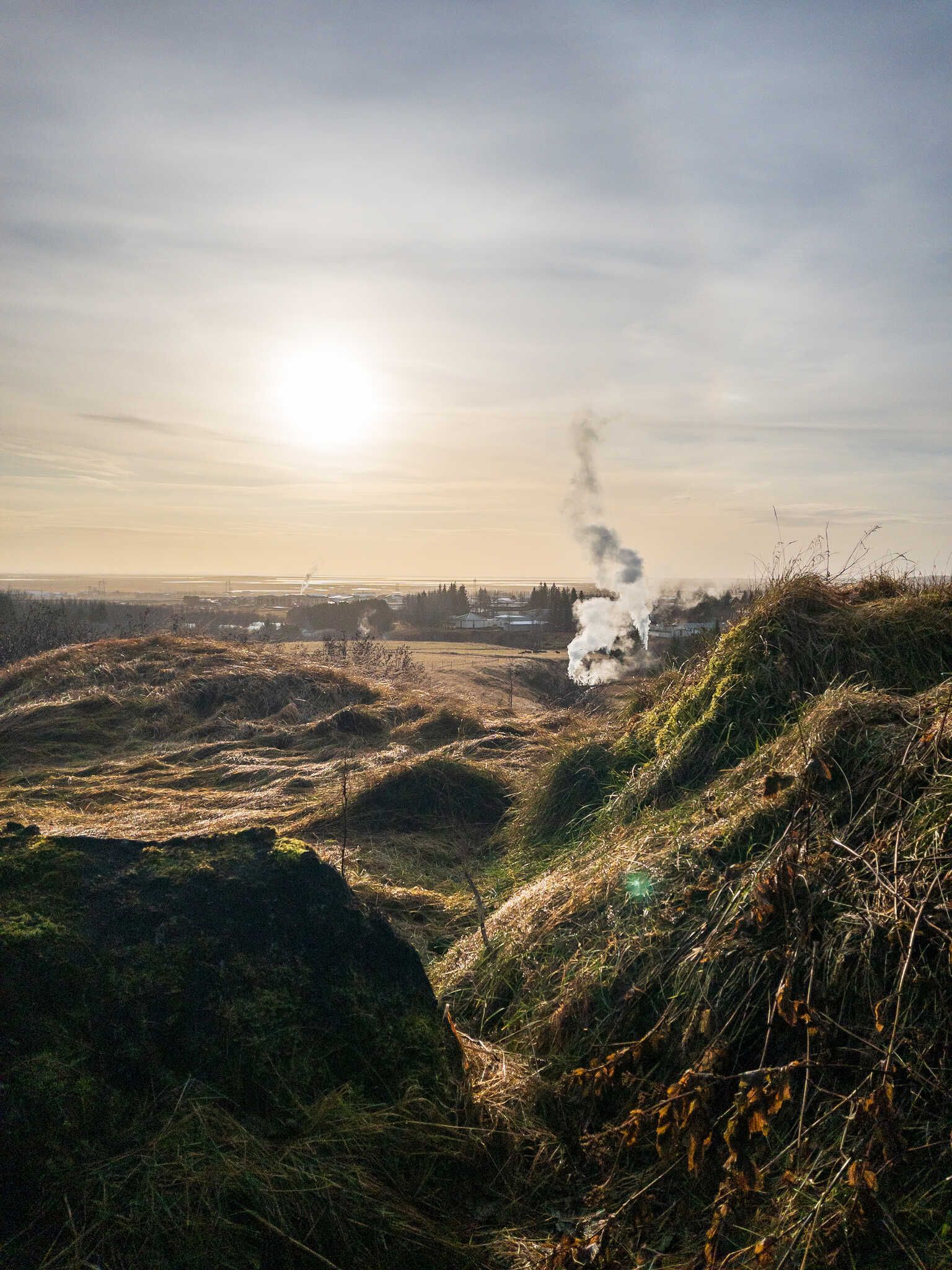 A view down a lumpy hillside. In the distance we see the town Hveragerði and the steam plume of a geothermal well.