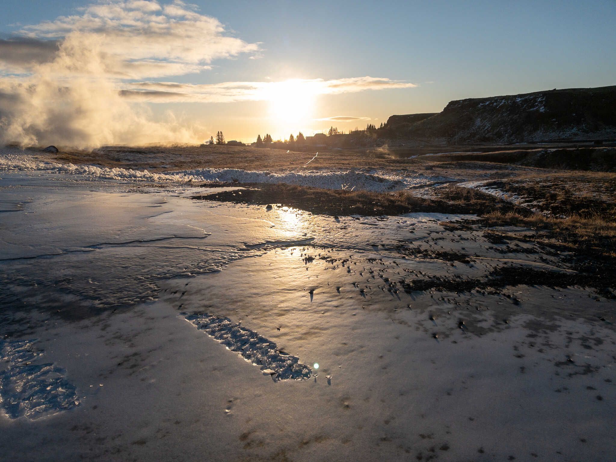 The steam from a geothermal well has left a shiny sheet of ice that reflects the sun. In the distance we see the silhouette of a treeline