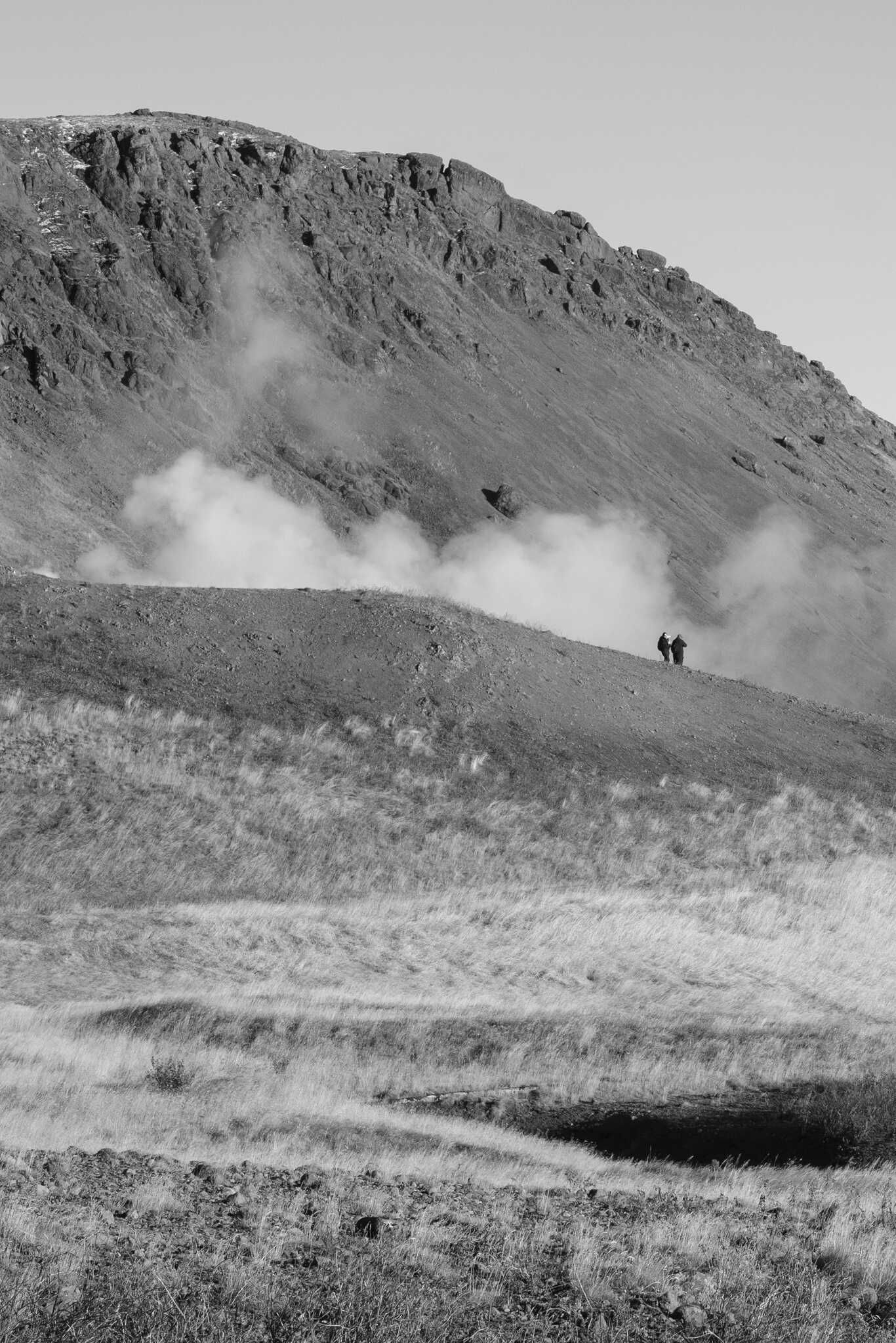 A couple of tourists walk along a hill. A cloud of steam rises in the background